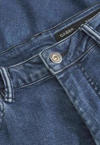 Modre kavbojkaste hlače z rahlim bledežem, srebrnim gumbom za zapiranje in črno tekstilno oznako z napisom "GABBA" blizu pasu.