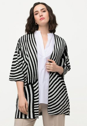 Zwart-witte gestreepte cardigan met korte mouwen, een open voorkant en een ruime pasvorm, gedragen over een wit shirt en beige broek.