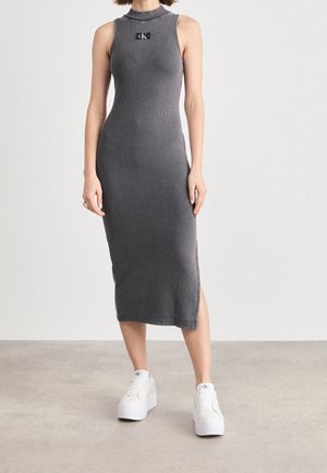 Szara sukienka na ramiączkach z prążkowanego materiału, o wysokim dekolcie i bocznym rozcięciu; ozdobiona logo Calvin Klein. Modelka ma na sobie białe sneakersy na platformie.