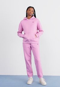 Haut à capuche en polaire rose avec poche kangourou et joggings assortis. Présente un petit logo sur le haut de la poitrine et à la cheville. Texture lisse.
