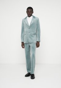 Fato de veludo em verde menta suave com colarinho picado, dois botões e bolsos laterais; combinado com uma camisola branca riscada e sapatos pretos.