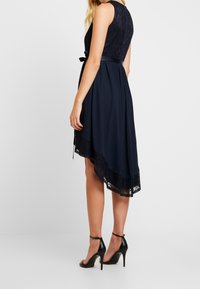 Robe bleu marine avec un corsage en dentelle, un ourlet asymétrique et une ceinture en satin. L'ourlet est orné d'une finition en dentelle, associé à des chaussures à talons hauts noires.