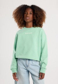 Felpa verde menta con polsini a costine, colletto a girocollo e la scritta "LA FINE DELL'ESTATE" in bianco sul petto. Indossata con jeans azzurri.