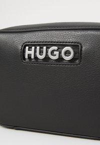 Svart skinnveske med sydd tekstur. Sølv "HUGO" merkevare på en rektangulær lapp. Glatte kanter og minimalistisk design.