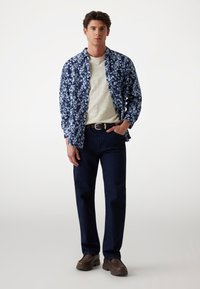 Camicia a maniche corte blu con motivo floreale sopra una t-shirt color crema, abbinata a jeans in denim scuro e scarpe slip-on marroni. Design semplice, vestibilità rilassata.