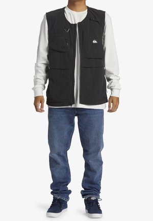 Man draagt een zwart werkvest met meerdere zakken over een wit shirt met lange mouwen, blauwe jeans en marineblauwe sneakers, staat rechtop.