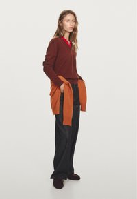 Maglione marrone sopra una camicia con colletto rosso, abbinato a jeans scuri larghi. Un cardigan arancione legato in vita aggiunge un colore di contrasto.