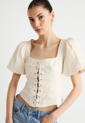 Elevandililla värvi blus pitsilise lukuga esiküljel, ruudukujulise kaelusega ja volangidega varrukatega. Kangas on tekstuuriga, silmahõlmadega kaunistatud ja vormis lõikega.