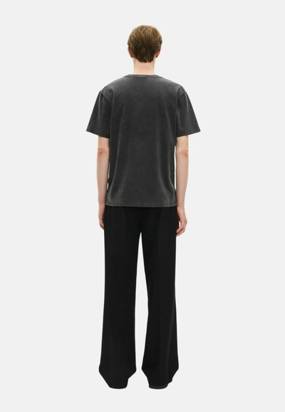 Persona de pie de espaldas, vistiendo una camiseta de manga corta negra desgastada y pantalones negros holgados contra un fondo blanco liso.