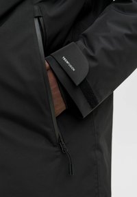 Črna nepremočljiva jakna z gladko teksturo, opremljena z zadrgo na žepu, Velcro zapestnicami in blagovno znamko "TECHFUSION" na rokavu.