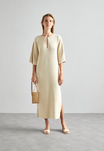 Γυναίκα φορώντας ένα κρεμ πλεκτό midi φόρεμα με άνοιγμα στον λαιμό και πλαϊνές σχισμές, κρατώντας μια μπεζ τσάντα και φορώντας κρεμ σανδάλια ανοιχτού σχεδίου.