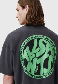 Mørkegrå oversized t-shirt lavet af bomuld, med et stort cirkulært grafisk motiv i grønt på ryggen, hvor teksten "ALLSAINTS" gentages.