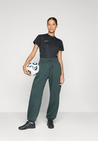Svart treningstrøye med hvit Nike-logo, grønne joggebukser med snøring, og svarte fotballsko. Holder en hvit fotball.