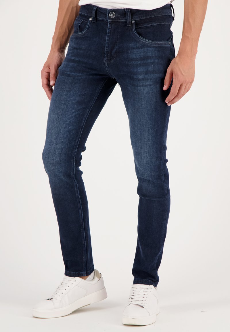Dark blue skinny jeans made of denim, featuring a classic five-pocket design, belt loops, and a button closure. Worn with white sneakers.