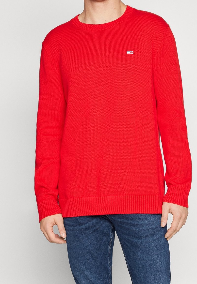 Roter gestrickter Pullover mit Rundhalsausschnitt, gerippten Bündchen und Saum. Kleines Logo-Detail auf der Brust. Kombiniert mit blauen Jeans.