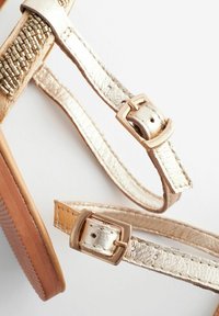Sandali metallici d'oro con cinturini intrecciati, fibbie regolabili e una suola texturizzata. Il design incorpora materiali lisci e texturizzati.