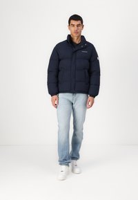 Navy pufferjack met een hoge kraag, rits aan de voorkant en twee zijzakken. Verloopt met lichtblauwe jeans en witte sneakers.