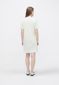 Svetlo bež bombažna obleka s kratkimi rokavi in sproščenim krojem, ki sega nad koleni. Gladek tekstura, preprost dizajn brez vidnih vzorcev.
