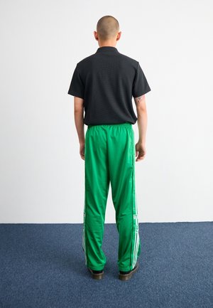 Persoon staat met de rug naar de camera, draagt een zwart shirt met korte mouwen, felgroene trainingsbroek met knopen aan de zijkant en zwarte schoenen op een blauw tapijt.
