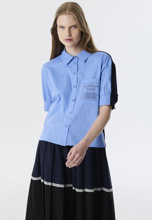 Camicia botton-down azzurro chiaro con maniche corte a palloncino e tasca sul petto con testo stampato. Abbinate a una gonna a pieghe blu navy e nera.