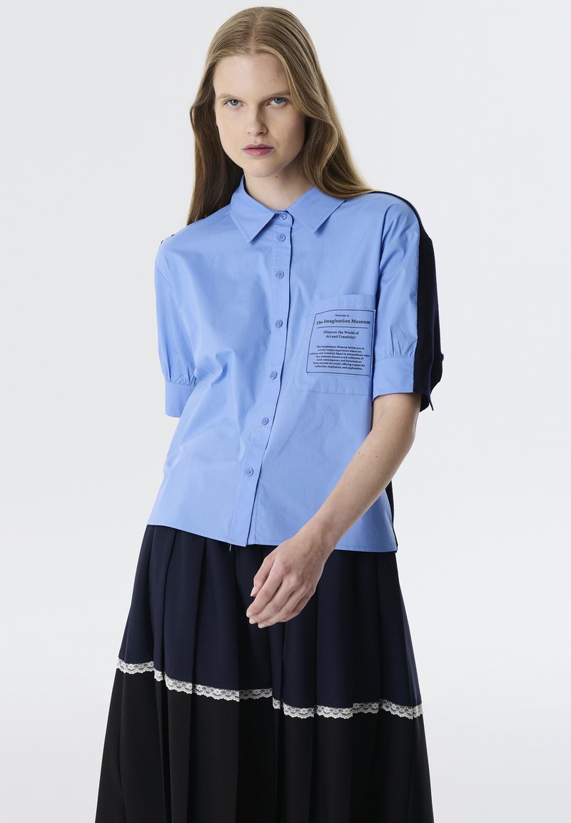 Camicia botton-down azzurro chiaro con maniche corte a palloncino e tasca sul petto con testo stampato. Abbinate a una gonna a pieghe blu navy e nera.