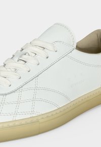 Baskets en cuir blanc avec semelle beige, lacets blancs, coutures détaillées et nom de la marque embossé sur le côté.