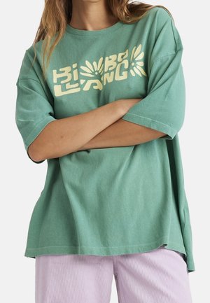Zielony oversize'owy t-shirt wykonany z bawełny. Zawiera wyrazisty żółty graficzny napis. Krótkie rękawy i luźny krój. Nałożony na jasnofioletowe spodnie.
