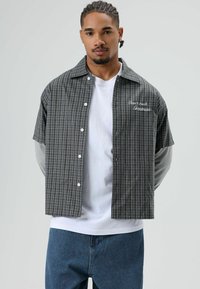 Kurzärmeliges kariertes Hemd in Grau und Schwarz mit weißen Knöpfen, versehen mit einem gestickten Spruch auf der Brust und darüber getragenem weißen Langarmshirt.