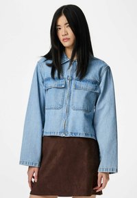 Jauna moteris su ilgais juodais plaukais dėvi šviesiai mėlyną trumpą džinsinį švarką ant rudos spalvos sijono, stovinti prieš baltą foną.