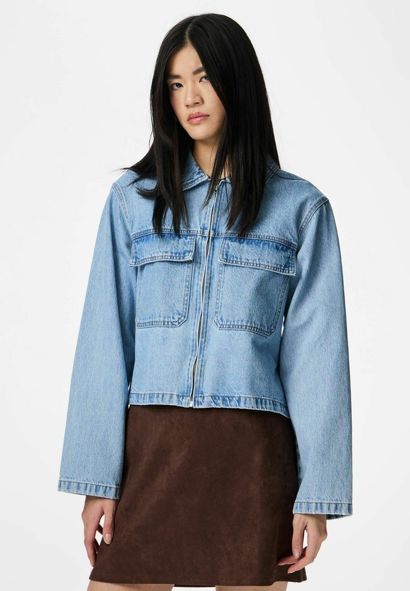 Jauna moteris su ilgais juodais plaukais dėvi šviesiai mėlyną trumpą džinsinį švarką ant rudos spalvos sijono, stovinti prieš baltą foną.