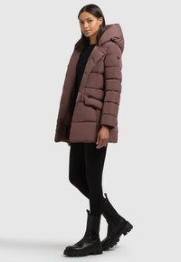 Egy nő barna párnázott kapucnis kabátot visel fekete felső, fekete leggings és fekete vastag talpú csizma felett, egy sima háttér előtt áll.