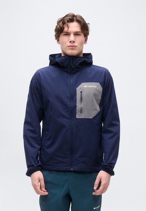 Jeune homme portant une veste Columbia bleu marine avec une poche poitrine grise et une capuche, debout devant un fond clair uni.