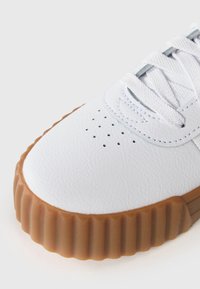 White leather sneaker with a textured surface, featuring a rubberized brown sole and perforated detailing near the toe. White laces included.