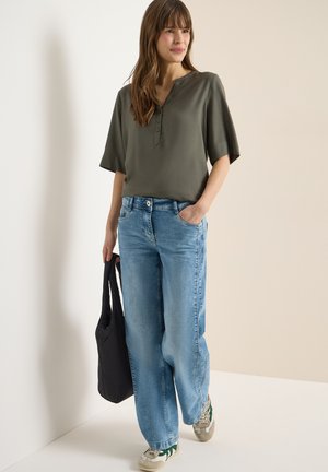 Femme debout contre un mur portant un chemisier olive à manches courtes, un jean large bleu clair, des baskets, et tenant un sac noir dans une main.