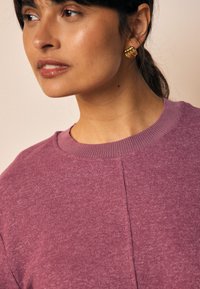 Pull violet côtelé avec col rond et détails de coutures apparentes, associé à des boucles d'oreilles créoles spirales dorées. Texture douce, design décontracté.