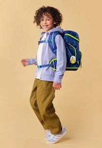 Blue backpack with geometric patterns and neon green accents. Child wearing a light purple hoodie and olive green pants, smiling.