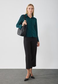 Donna con una blusa lunga a maniche lunghe color teal, pantaloni neri alla caviglia e scarpe con tacco nere, che porta a spalla una borsa nera texturizzata.