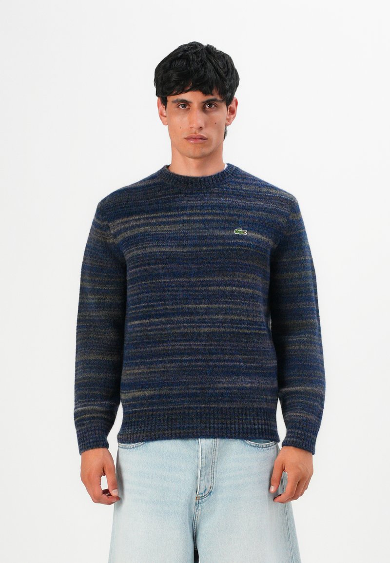 Uomo che indossa un maglione a righe blu e grigie con il logo Lacoste e pantaloni corti in denim azzurro, in piedi contro uno sfondo bianco uniforme.