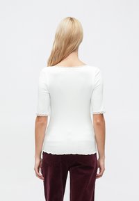 Valkoinen lyhythihainen paita, jossa aaltoileva pääntie ja pitsireunus hihoissa, valmistettu ribbineuloksesta. Kuvattu takaa.