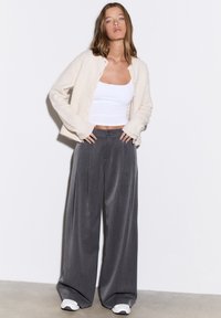 Kardigan in Cremeton, weißes Tanktop und graue, weit geschnittene Hose mit strukturierter Passform; weiche Textur und feine Nadelstreifen; weiße Turnschuhe.