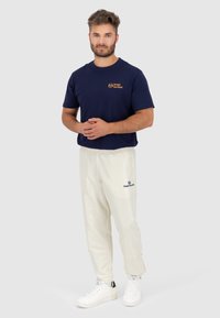 Marineblauwe t-shirt met oranje logo, crèmekleurige trainingsbroek en witte sneakers. De outfit heeft een ontspannen pasvorm en een gladde stoftextuur.