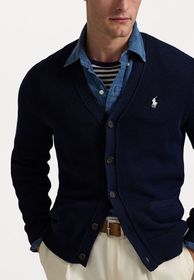Marinblått stickat kardigan med v-ringning, knapplist och två fickor, parat med en blå denimskjorta och randig undertröja.