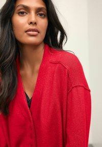 Cardigan rouge avec un col en V et une fermeture à boutons, mettant en avant un tricot texturé et un détail en tricot aérien aux épaules.