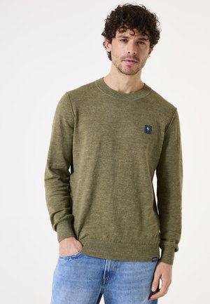 Olivengroen gebreide trui met ronde hals en geribbelde manchetten. Een klein logo-opdruk is op de linkerborst. Gecombineerd met blauwe spijkerbroek.