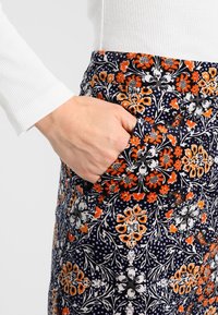 Main dans la poche d'une jupe à motif floral vibrant, portant un haut à manches longues côtelé blanc devant un fond uni.