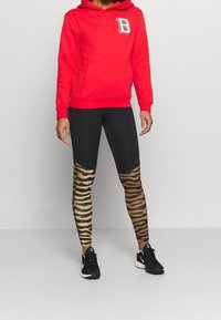 Czerwona bluza z kapturem z białą naszywką w literę "B", połączona z czarnymi legginsami w wyrazisty, złoty i czarny wzór w paski zebry. Czarne sneakersy.