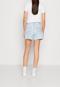 Mujer vestida con una camiseta blanca, pantalones cortos de denim azul claro, calcetines blancos y zapatillas Converse blancas, de pie de espaldas contra un fondo liso.