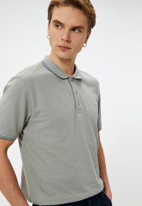 Grå polo-shirt lavet af tekstureret stof, med en klassisk krave, tre-knaps lukning og blå accentstriber på kraven og ærmerne.