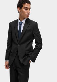 Uomo in completo nero e camicia bianca indossa una cravatta blu, con una mano in tasca, guardando in basso e di lato su uno sfondo semplice.