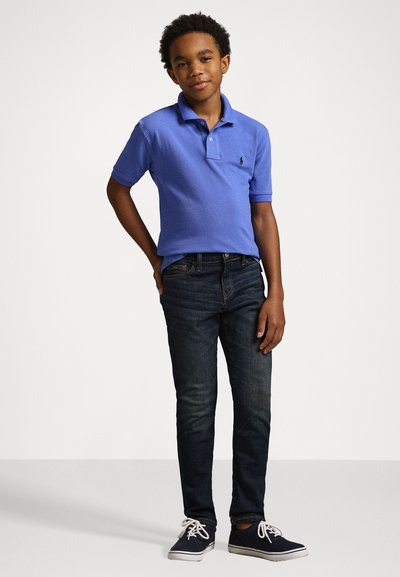 Camisa polo azul com uma pala de dois botões, jeans escuros e ténis azul-marinho. A camisa apresenta um pequeno logótipo no peito, em tecido de malha texturada.